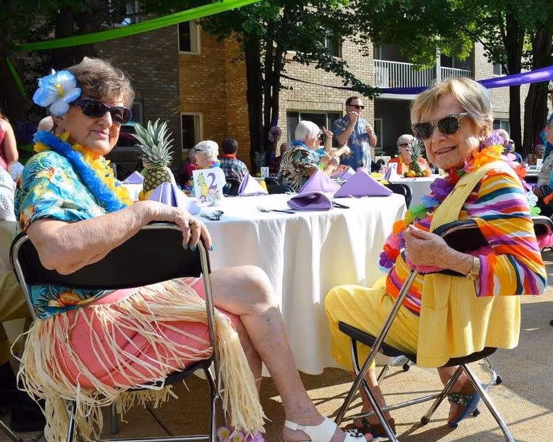 Two elderly women wearing sunglasses and colorful Hawaiian-themed clothing and accessories sit on folding chairs at an outdoor event with tables covered in white tablecloths and purple napkins. Other people are visible in the background near a brick building, and there are green and purple streamers hanging above.