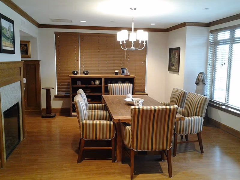 A dining room with a wooden table surrounded by six striped upholstered chairs. The room has wooden flooring, a fireplace on the left, a chandelier hanging above the table, and windows with blinds on the right and back walls. There are paintings on the walls and a bust sculpture near the window.