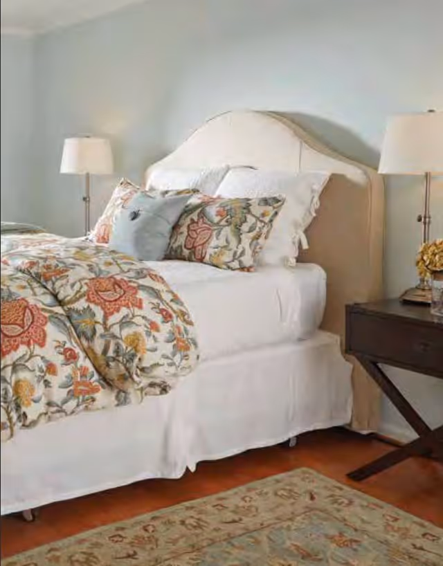 A neatly made bed with a beige upholstered headboard, white sheets, and a floral patterned comforter and pillows. There are two bedside tables, each with a lamp, and a decorative rug on the wooden floor.