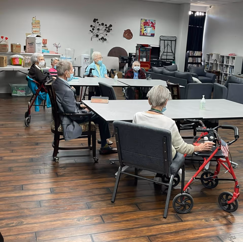 A group of elderly individuals wearing face masks sitting around tables in a spacious room with wooden flooring. Some use walkers and wheelchairs. The room has couches, bookshelves, and a popcorn machine in the background, suggesting a common area for socializing or activities.