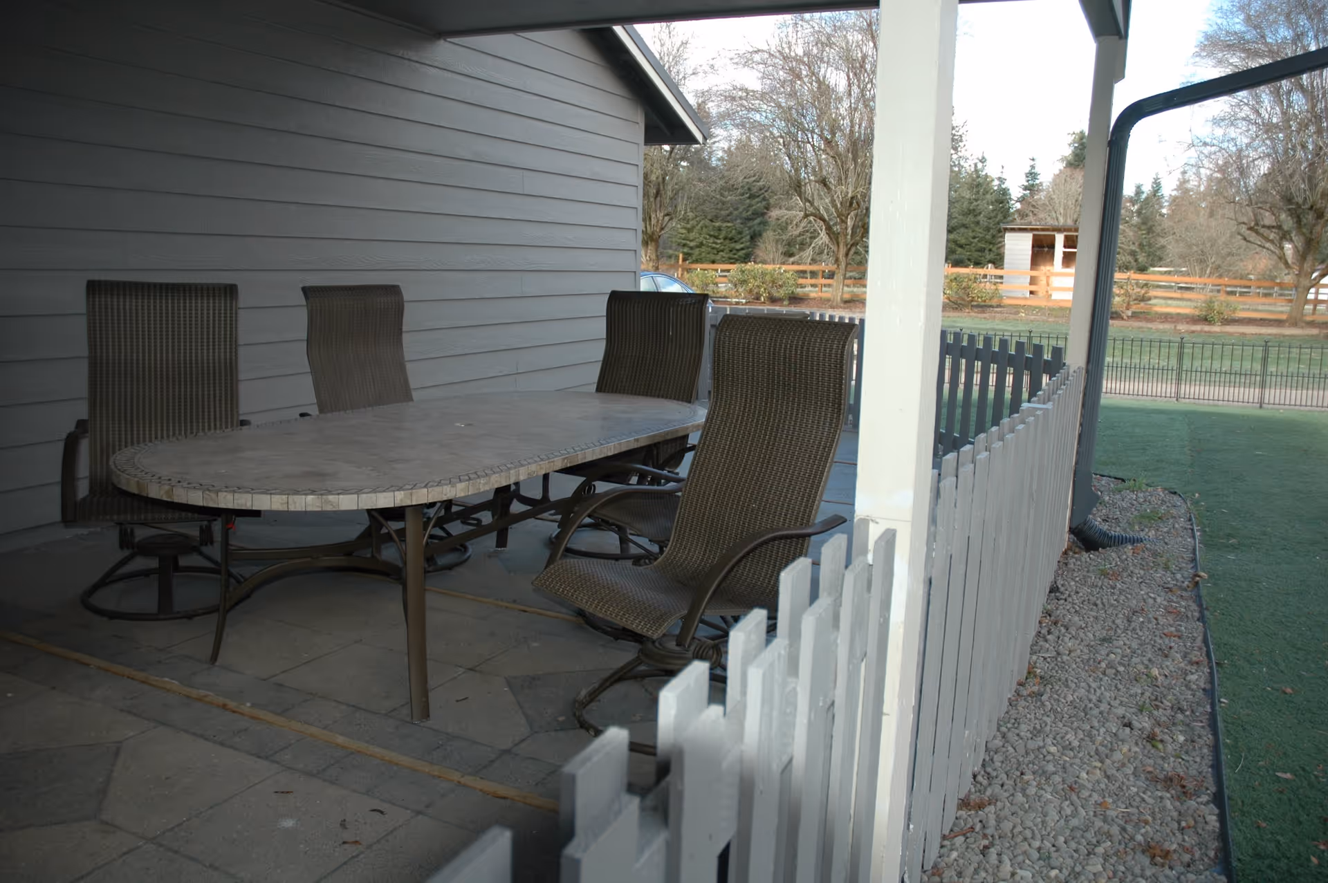 Covered outdoor patio with an oval table, several wicker chairs, and a white picket fence overlooking a lawn and trees.