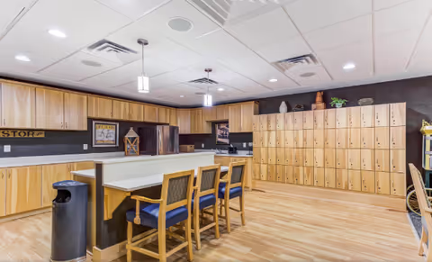 A spacious kitchen area with wooden cabinets, a refrigerator, and a countertop with three wooden chairs with blue cushions. The room has a light wood floor and a wall with multiple wooden lockers. There are pendant lights hanging from the ceiling and decorative items on top of the lockers.