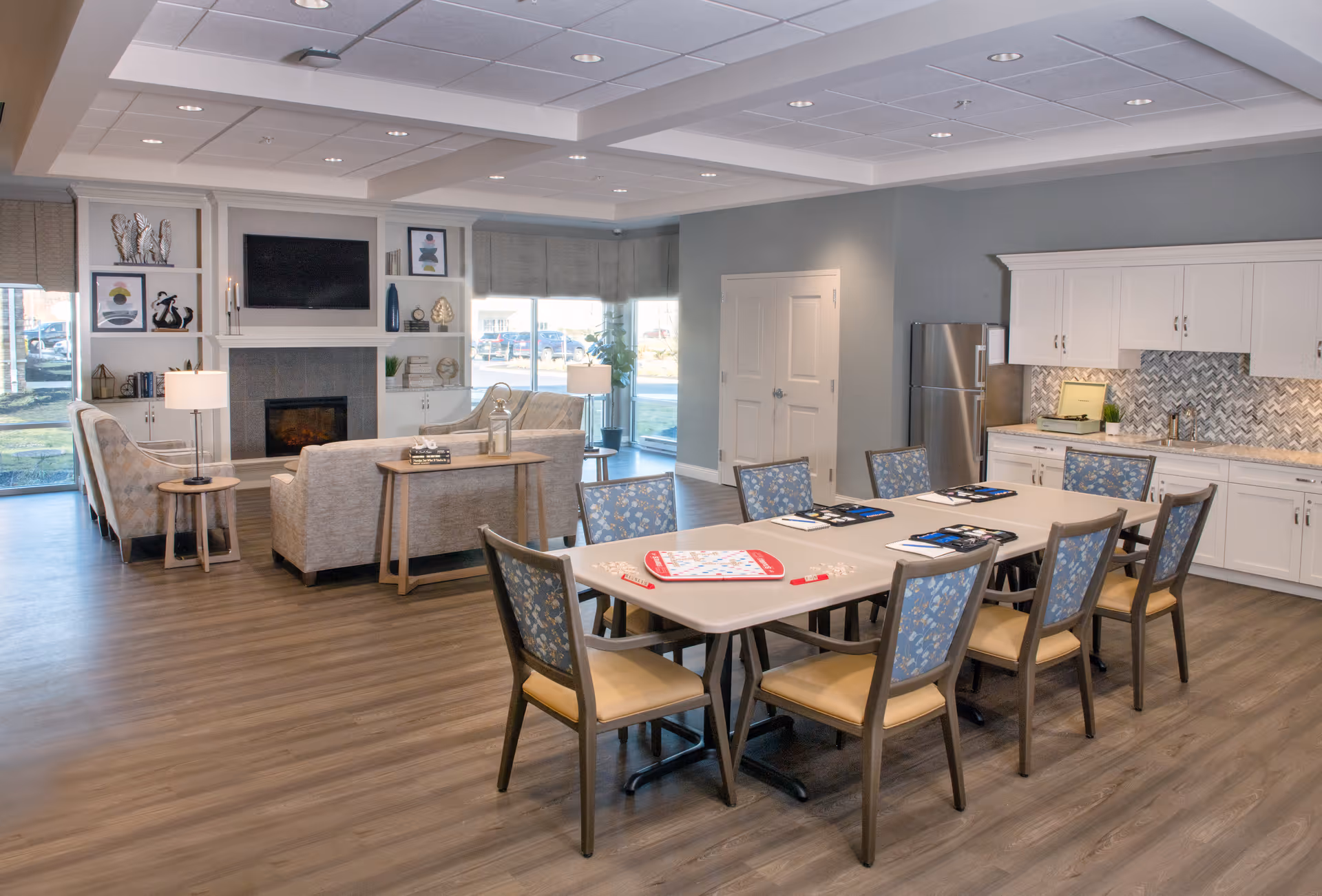 A spacious and well-lit common area in a senior living facility featuring a rectangular table with eight chairs arranged around it. On the table are game boards and booklets. In the background, there is a cozy seating area with two sofas, a fireplace, a wall-mounted TV, and built-in shelves with decorative items. To the right, there is a kitchenette with white cabinets, a stainless steel refrigerator, a sink, and a patterned backsplash. Large windows allow natural light to fill the room.
