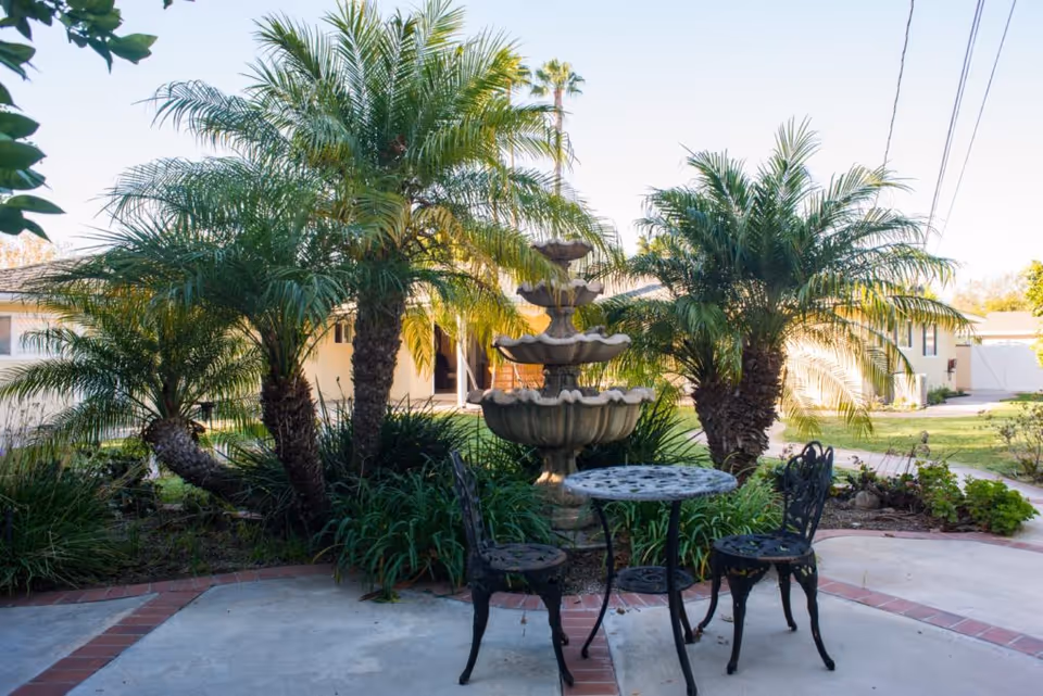 Outdoor patio area with a small round metal table and two matching chairs in front of a multi-tiered stone fountain, surrounded by palm trees and greenery, with a building visible in the background under a clear sky.