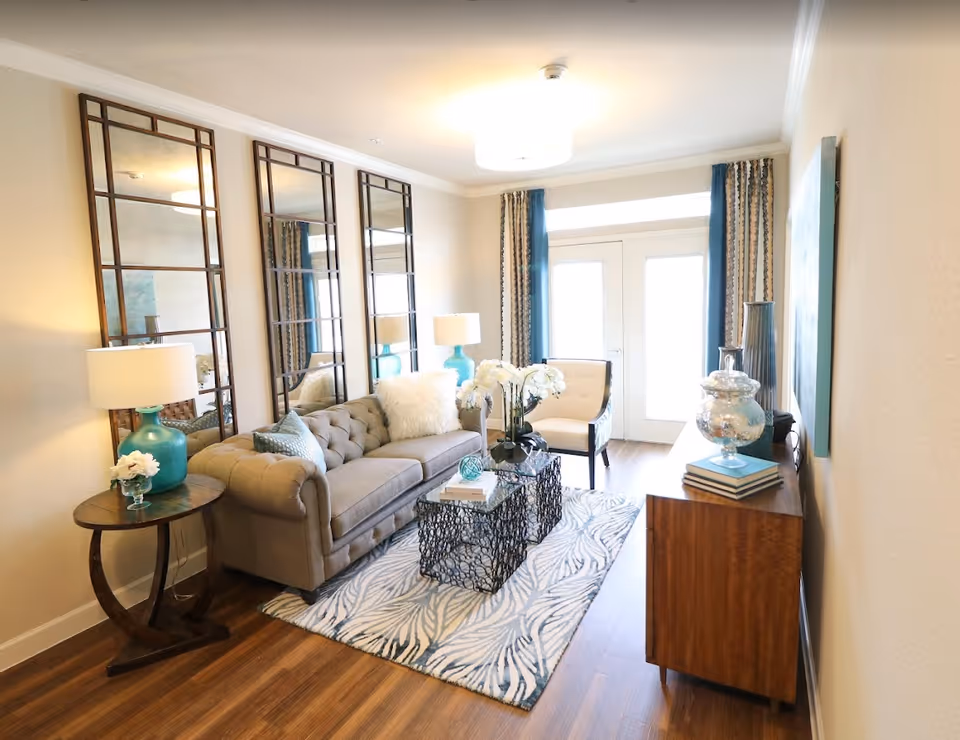 A bright and cozy living room with a beige tufted sofa adorned with pillows, a white armchair, and two decorative black metal coffee tables on a patterned rug. The room features three tall mirrors on one wall, two turquoise table lamps on either side of the sofa, a wooden side table with a small flower arrangement, and a wooden console table with decorative items. French doors with blue and patterned curtains allow natural light to fill the space.