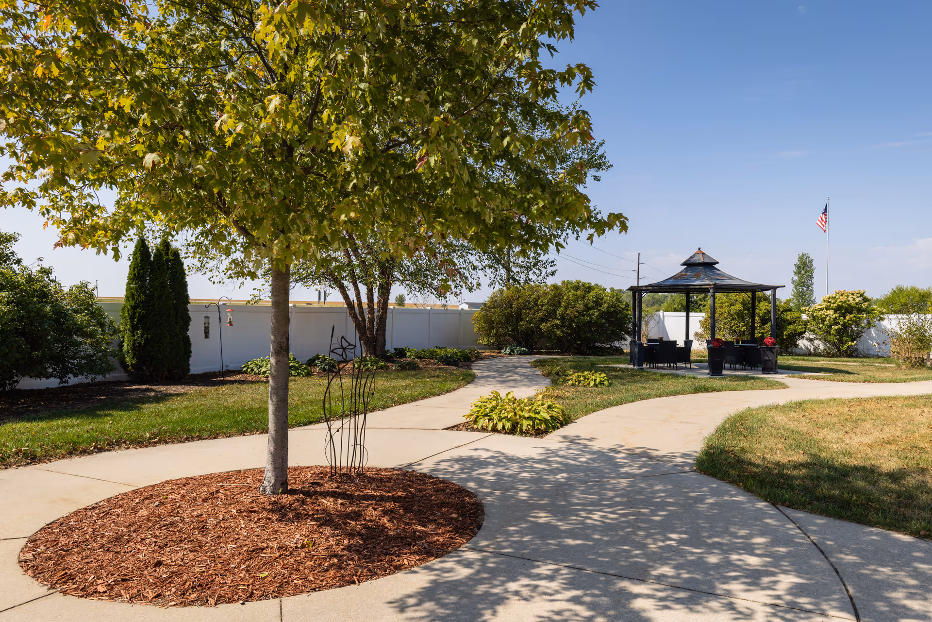 Landscaped courtyard with a tree, curving paved walkways and a gazebo under a clear blue sky.