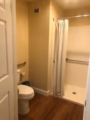 A bathroom with a white toilet, a toilet paper holder, a grab bar on the wall, a shower area with a white curtain, and wooden flooring.