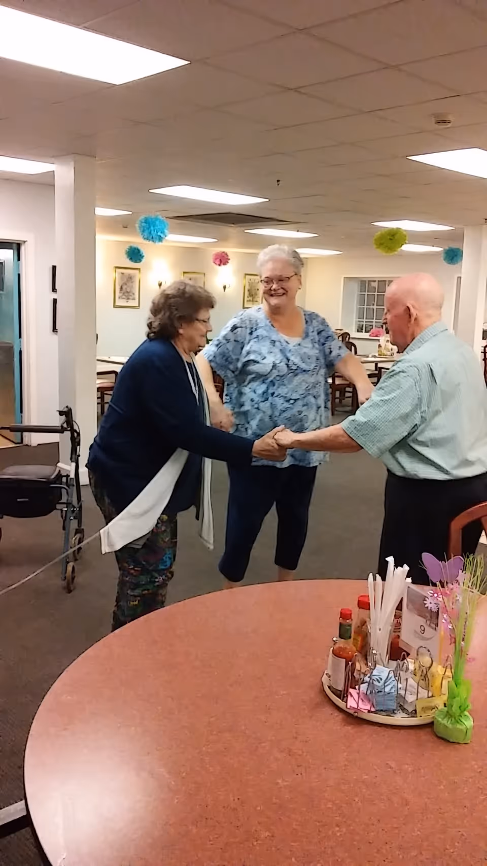 Three elderly residents holding hands and smiling while dancing in a decorated dining/activity room.