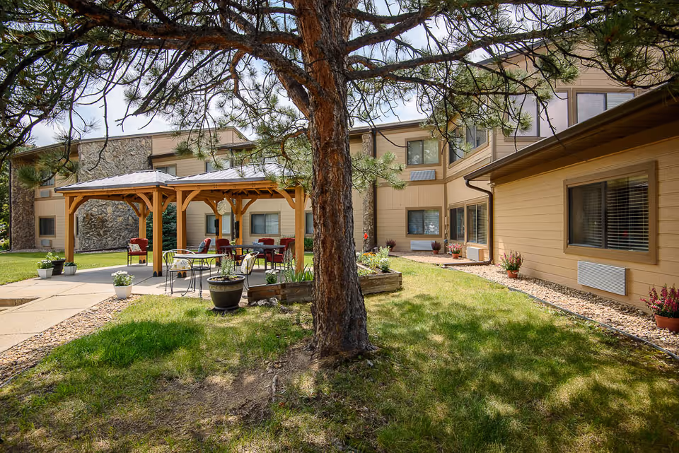Outdoor garden area at Life Care Center of Evergreen featuring a large tree in the foreground, a wooden gazebo with seating underneath, potted plants, and a two-story beige building with multiple windows in the background.