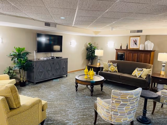 A cozy living room with a brown sofa adorned with patterned cushions, two patterned armchairs, a yellow armchair, a round wooden coffee table with decorative yellow items, a TV mounted on a dark wooden cabinet, and various lamps and plants providing warm lighting.