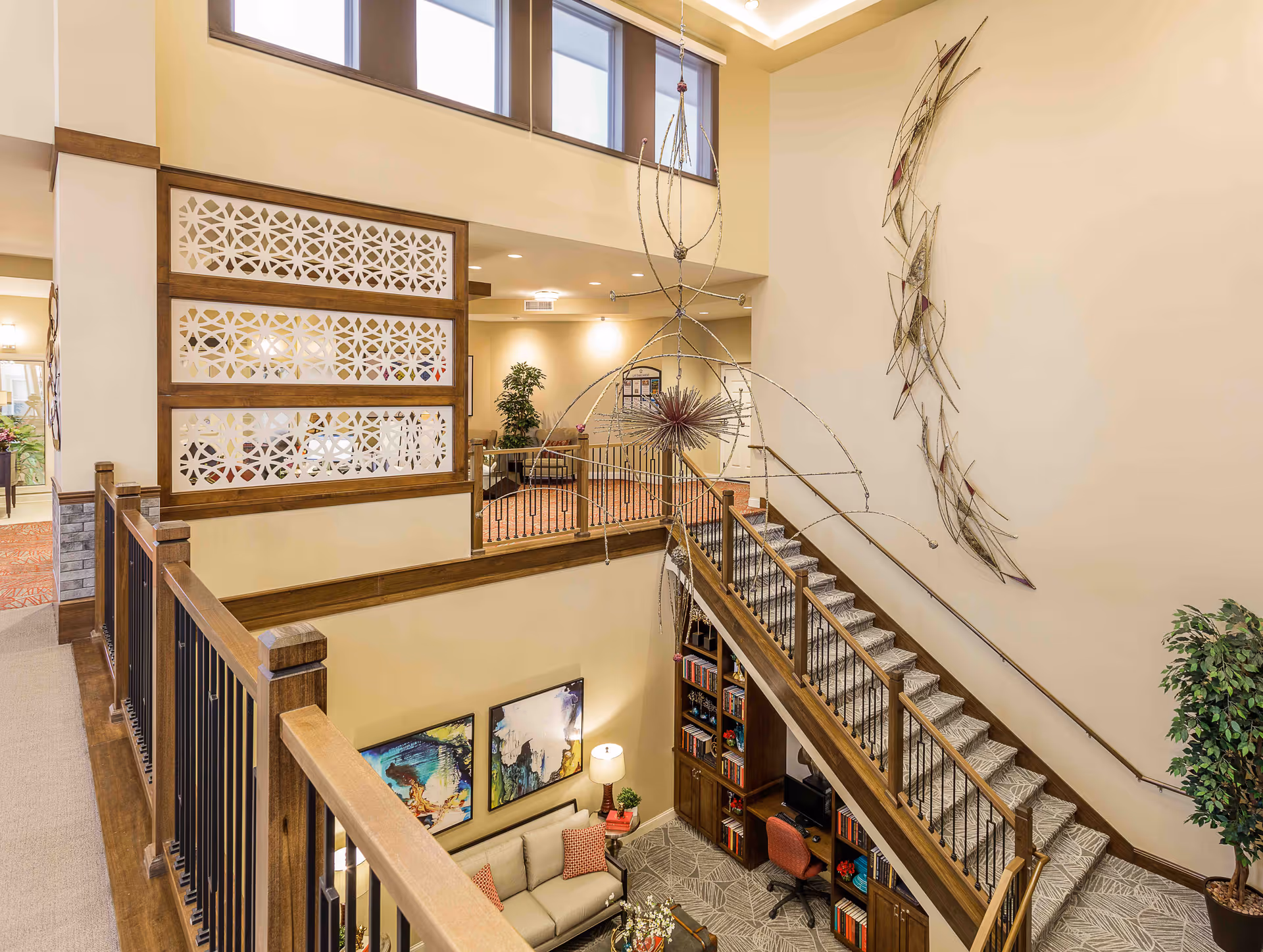 Interior view of a senior living facility showing a two-story common area with a staircase, decorative wall art, a seating area with a sofa and colorful pillows, bookshelves, and a desk with a chair. The space is well-lit with natural light from high windows and warm artificial lighting.