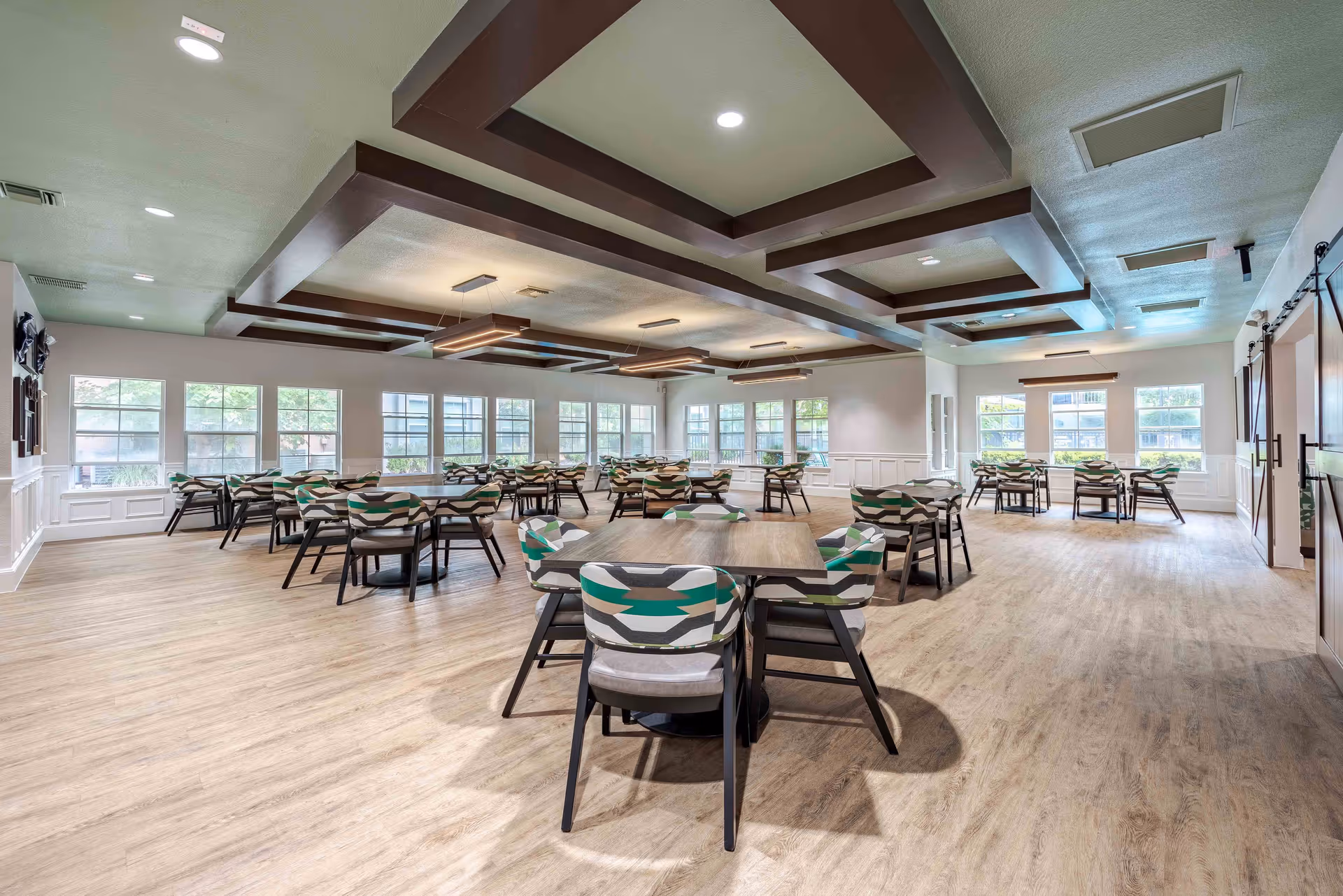 A spacious dining room with multiple tables and chairs arranged neatly. The chairs have patterned upholstery in shades of green, gray, and white. Large windows line the walls, allowing natural light to fill the room. The ceiling features decorative wooden beams with modern rectangular light fixtures hanging down. The floor is covered with light wood-style flooring.