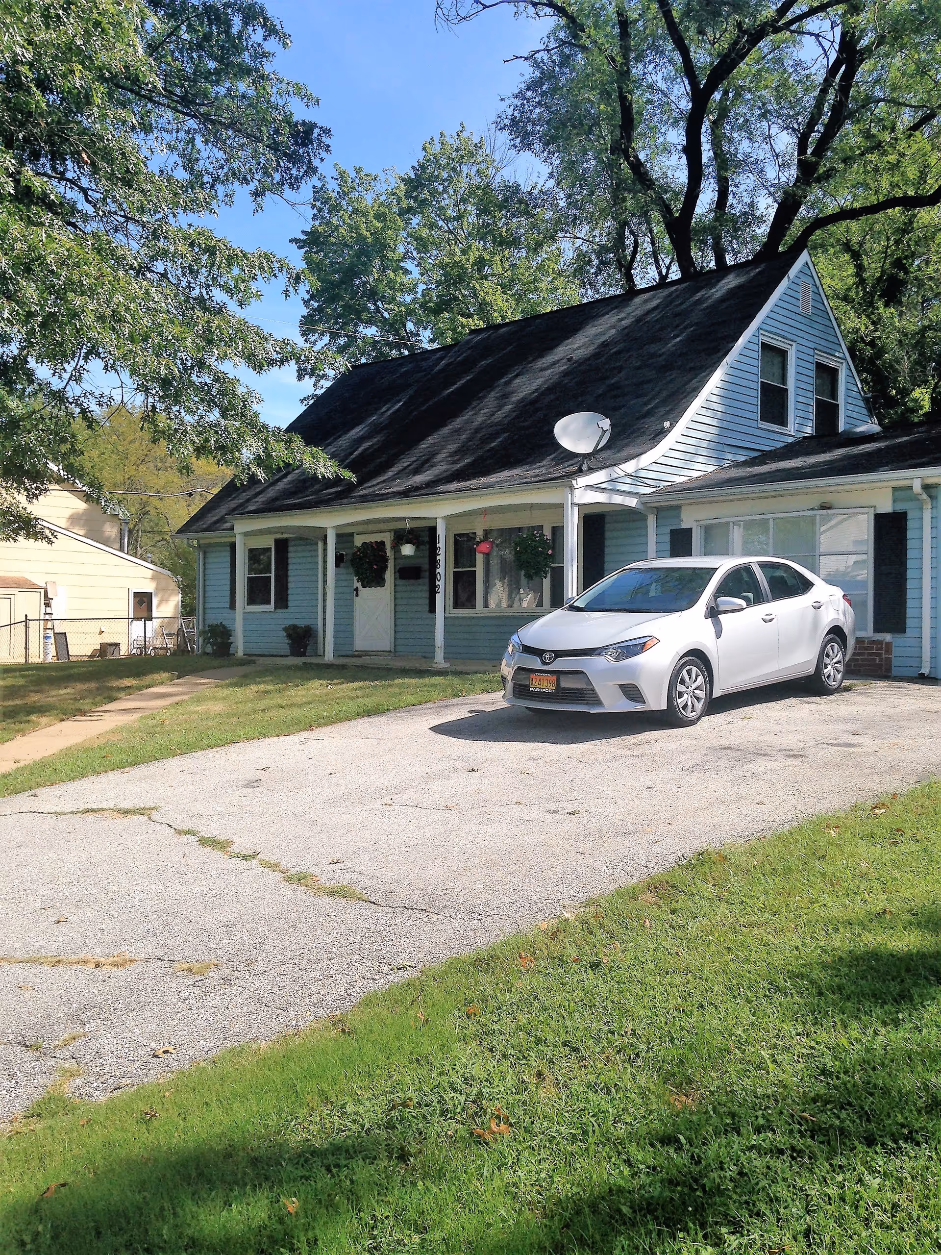 A light blue house with a dark roof and white trim, featuring a front porch with hanging flower baskets. A silver sedan is parked in the driveway. The house is surrounded by green grass and trees under a clear blue sky.