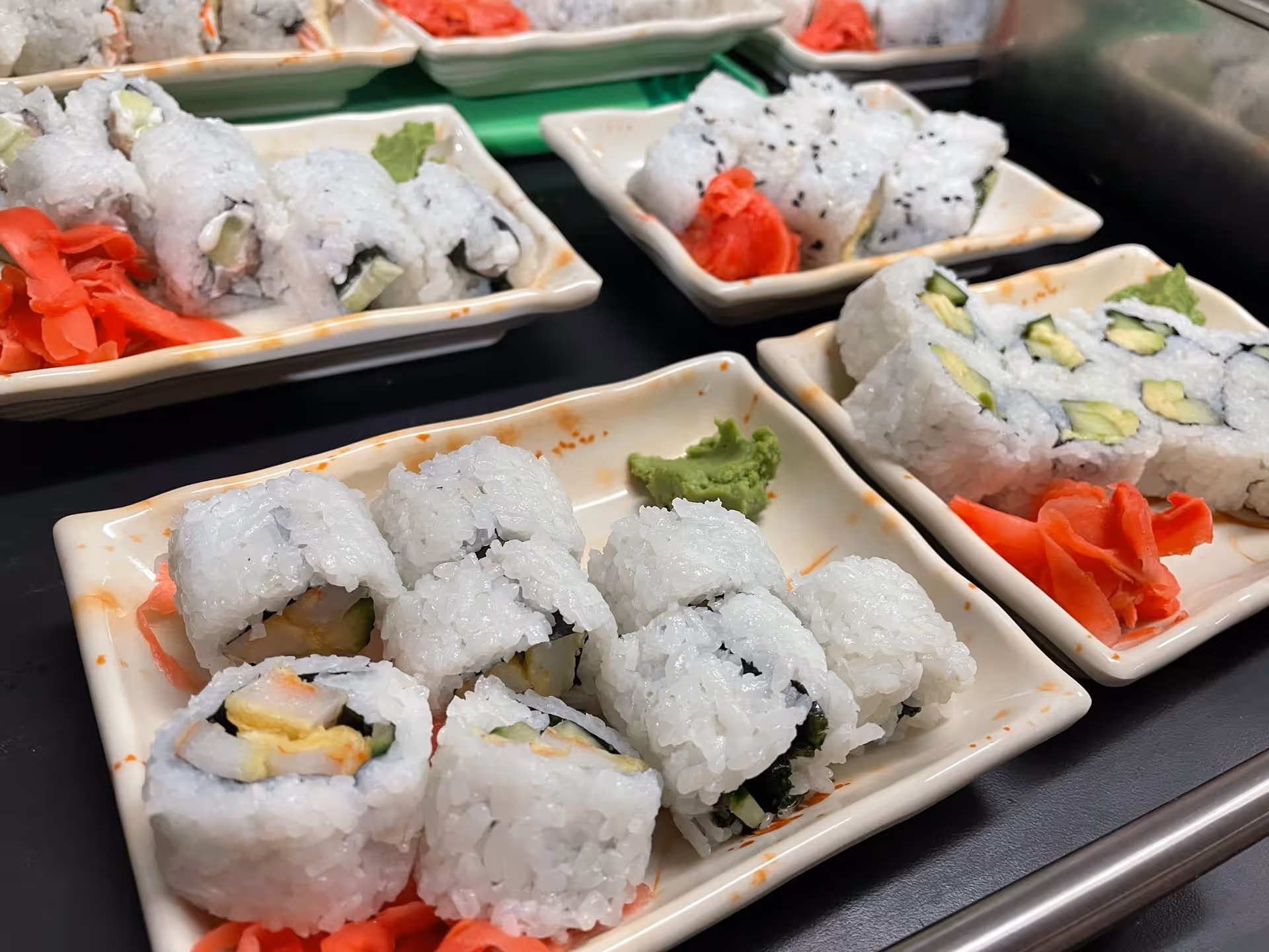 Close-up view of several small rectangular plates with sushi rolls, garnished with pickled ginger and wasabi, arranged on a black surface.