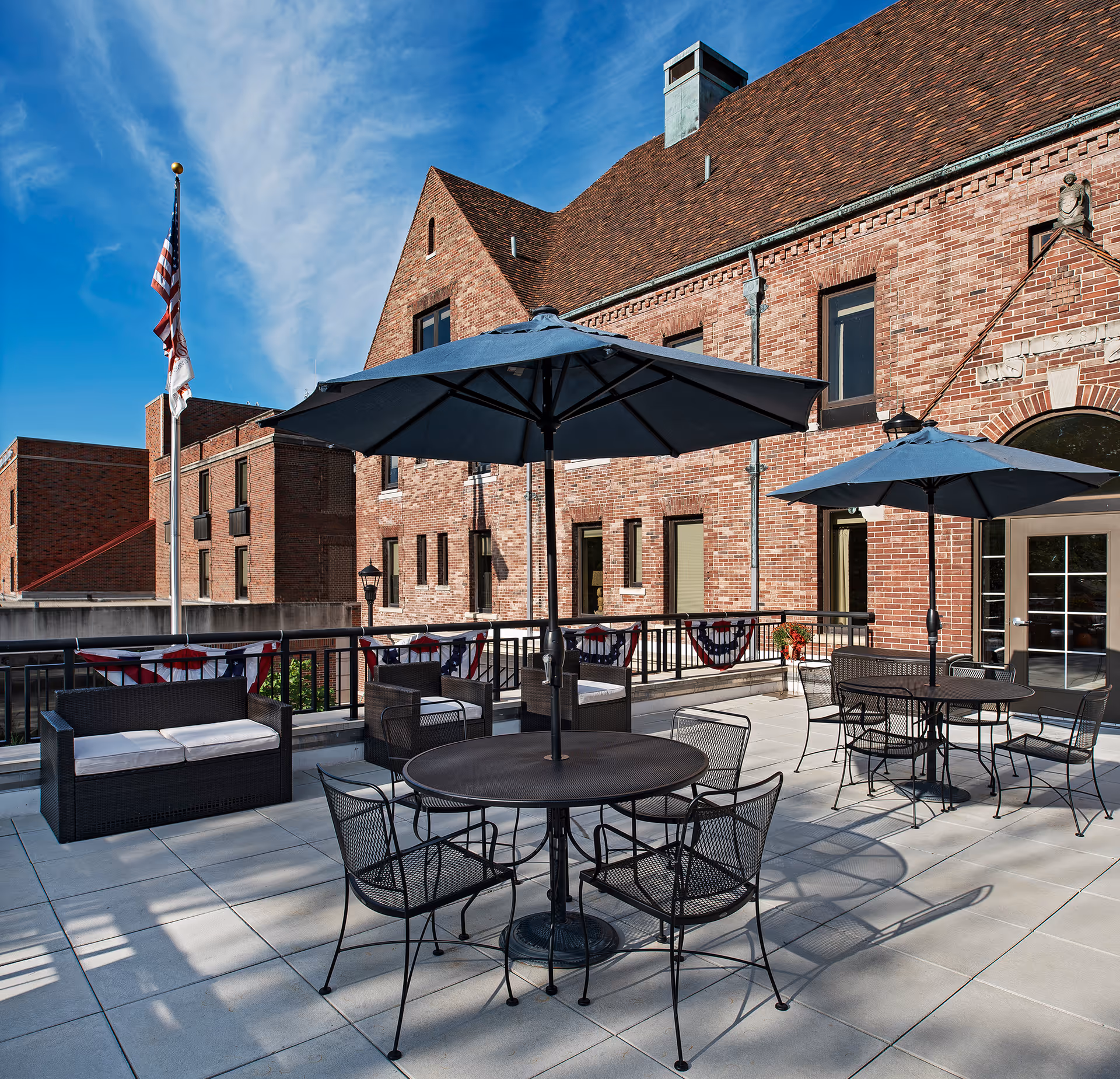 Outdoor patio area with black metal tables and chairs, each table shaded by a large blue umbrella. There is a cushioned black wicker loveseat on the left side. The patio is adjacent to a large brick building with multiple windows and a door. An American flag and another flag are flying on a flagpole to the left. The sky is clear and blue.