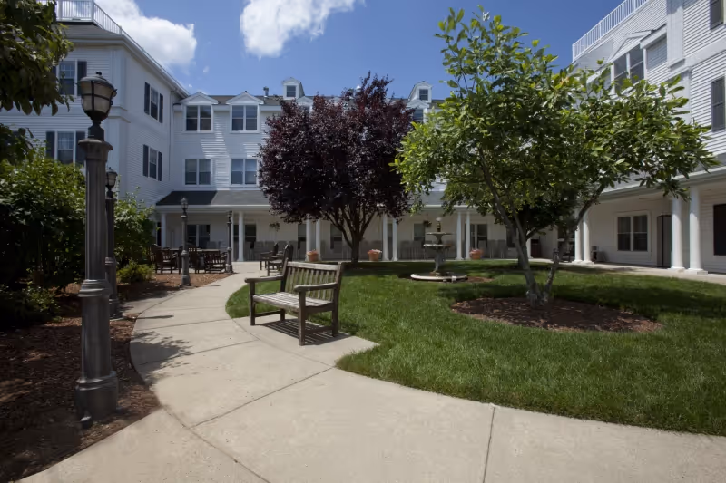 Outdoor courtyard area at Whitney Place at Westborough featuring a curved concrete walkway, wooden benches, green grass, trees, and a multi-story white building with windows and columns surrounding the courtyard.