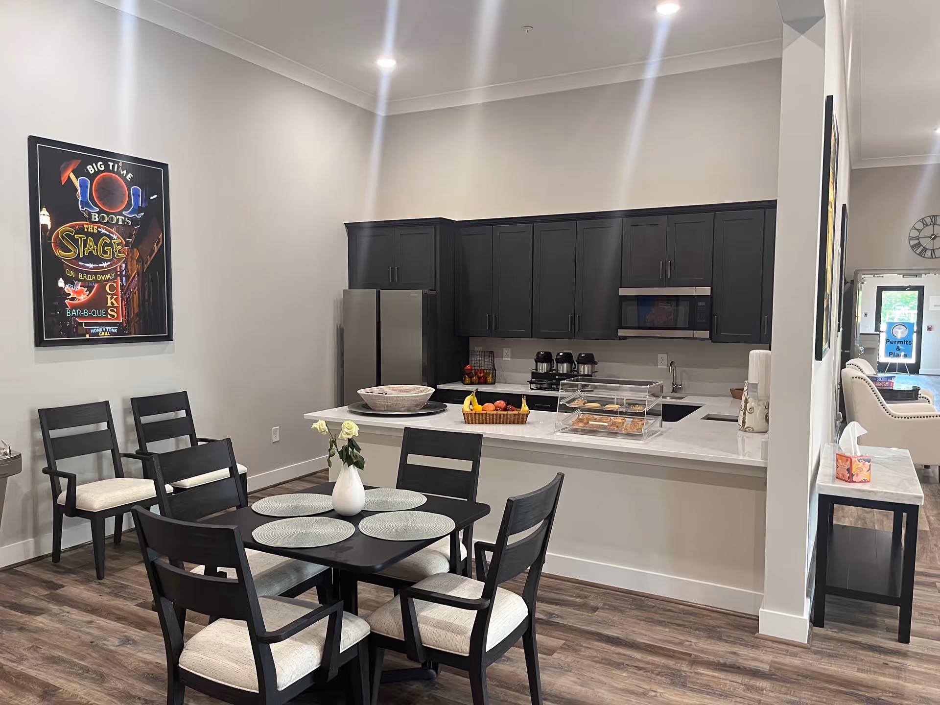 A modern kitchen and dining area with dark cabinets, a stainless steel refrigerator, microwave, and coffee makers. A black dining table with four chairs is set with placemats and a white vase with flowers. A framed colorful poster hangs on the wall, and a countertop displays a basket of fruit and a clear container with pastries. The flooring is wood, and the space is well-lit with ceiling lights.