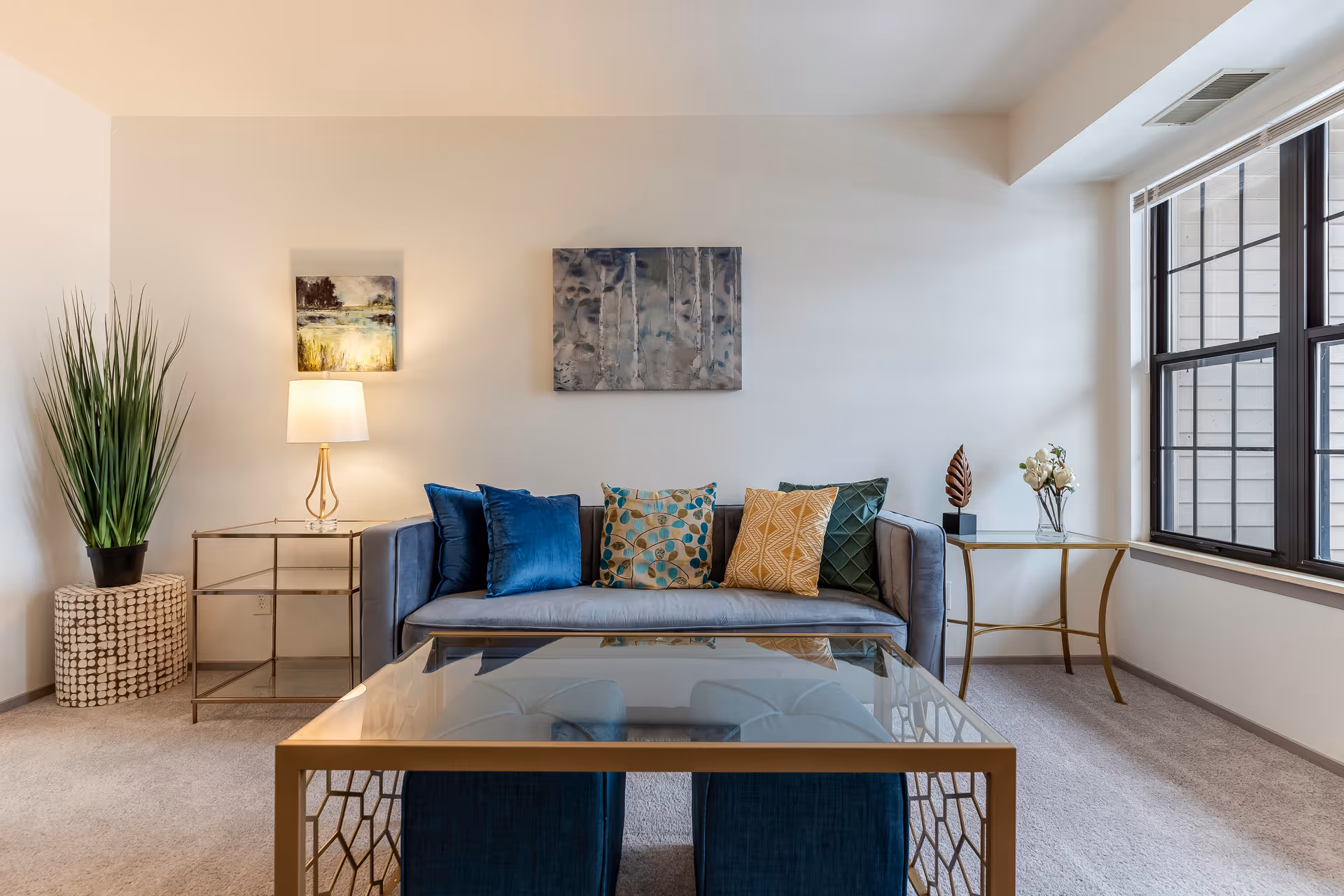 A modern living room with a gray sofa adorned with colorful throw pillows in blue, green, and yellow patterns. There is a glass coffee table with a gold frame in front of the sofa. To the left, a side table with a lamp and a tall green plant in a decorative pot. On the right, a small gold-framed side table with a vase of white flowers and a decorative leaf sculpture. Large windows with black frames allow natural light to fill the room. Two pieces of artwork hang on the wall above the sofa.