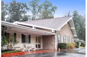Front exterior of a single-story brick senior care building with a covered entrance, benches, landscaping, and wet pavement.