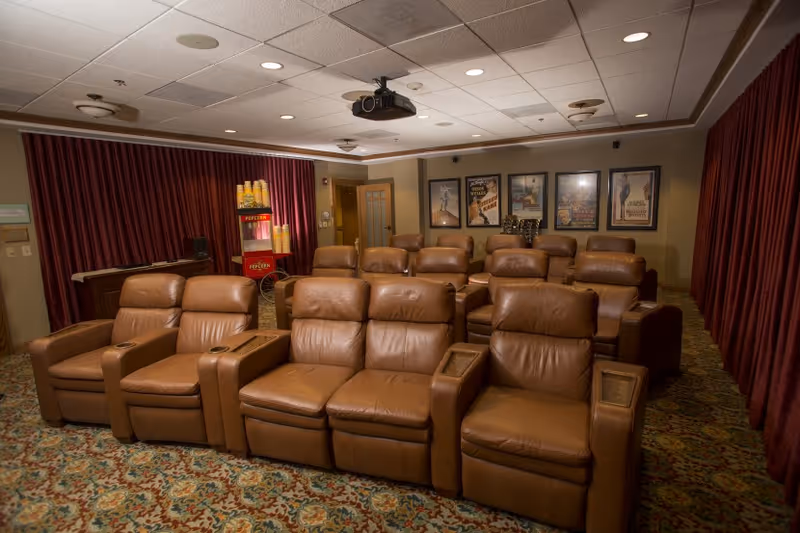 A cozy movie theater room with three rows of brown leather recliner seats facing a projector. The room has patterned carpet, red curtains on the side walls, and framed movie posters on the back wall. A popcorn machine is visible in the back corner near the entrance.