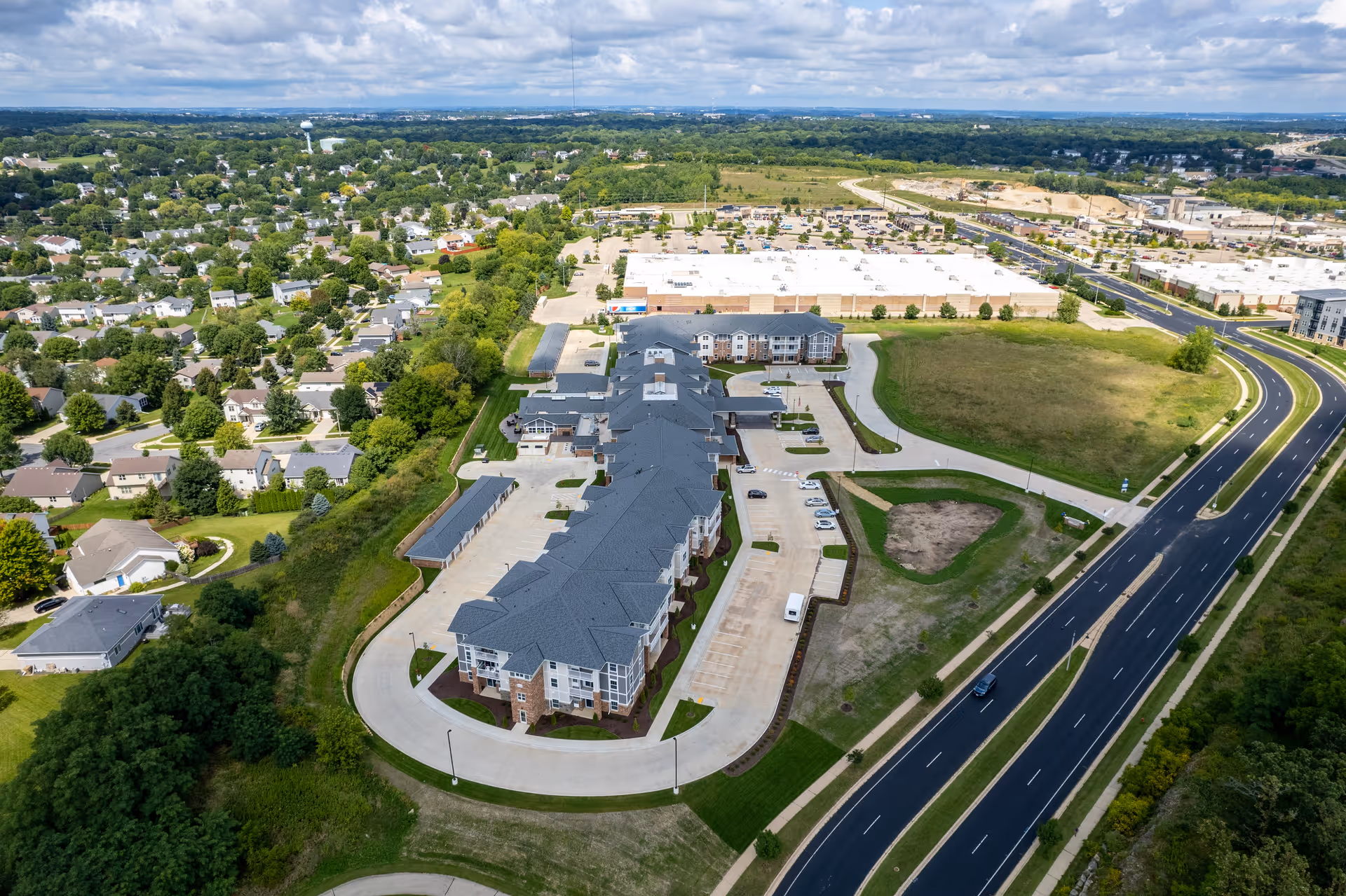 Aerial view of Quarry Ridge Retirement Community showing a large multi-building complex with parking lots, surrounded by green spaces, residential neighborhoods, and a nearby highway under a partly cloudy sky.