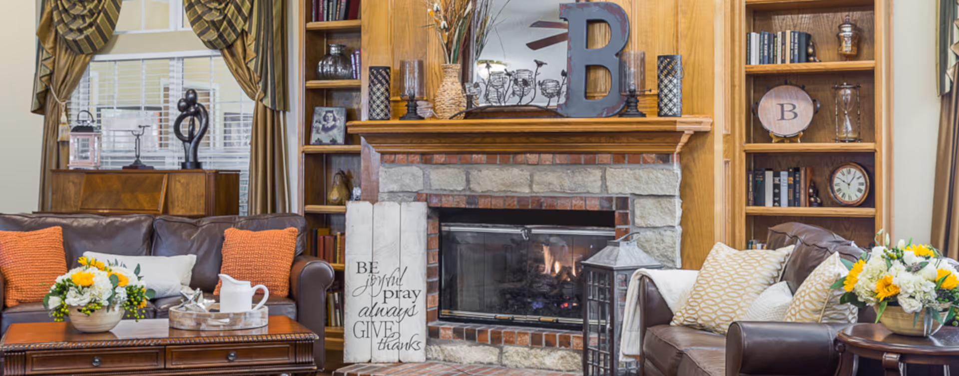 Cozy living room with two brown leather sofas adorned with orange and patterned pillows, a wooden coffee table with flower arrangements and a tray, a stone fireplace with a wooden mantle decorated with a large letter B, vases, and framed photos, flanked by built-in wooden bookshelves filled with books and decorative items.