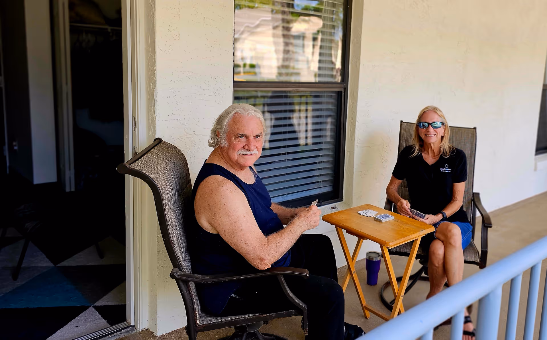 Two older adults sitting on a covered porch playing cards at a small folding table.