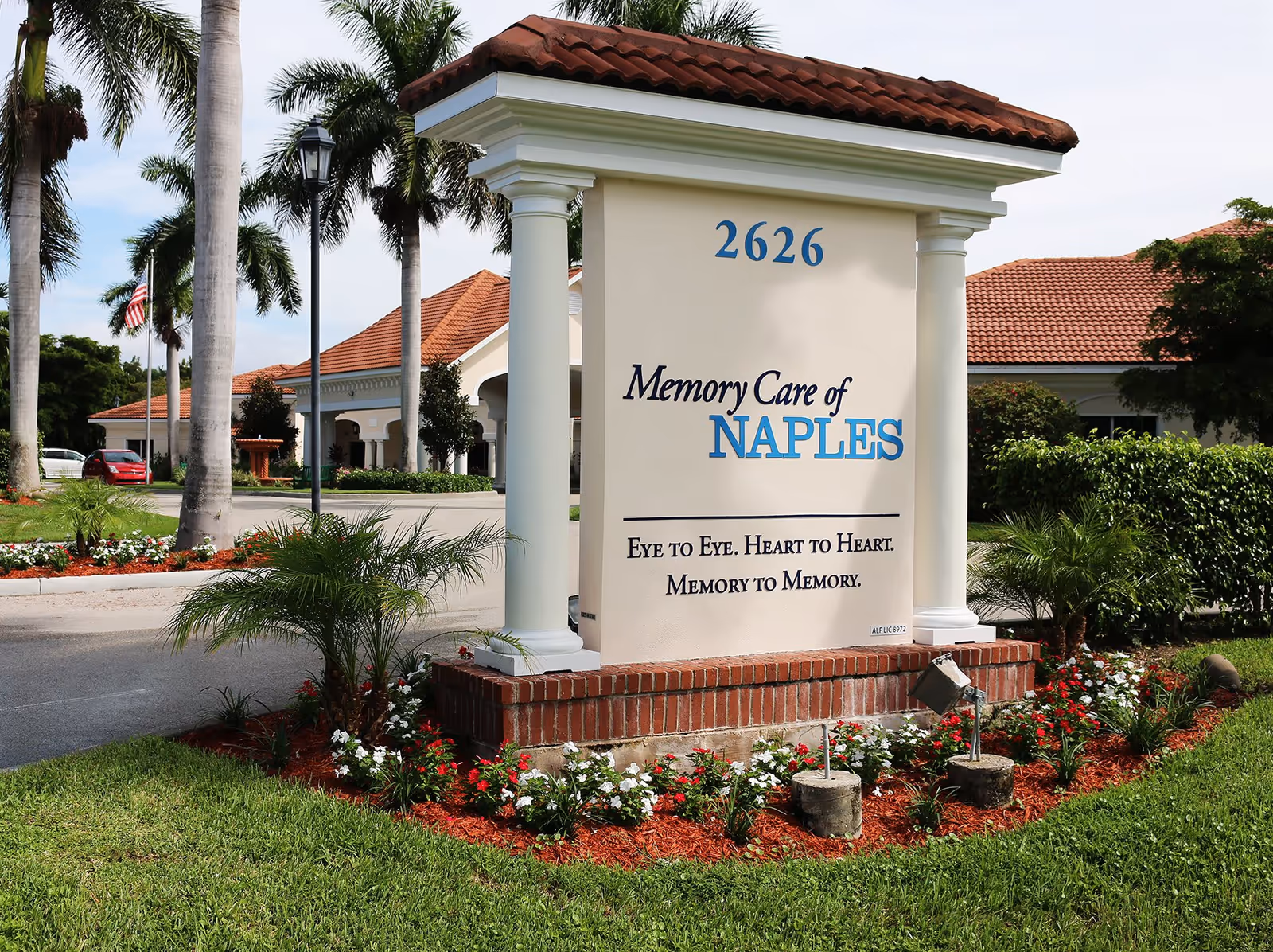 Outdoor view of a sign for Memory Care of Naples at 2626, surrounded by palm trees, flowers, and well-maintained landscaping with a building featuring a red tile roof in the background.