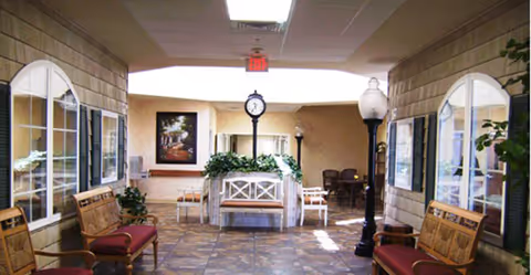 Indoor hallway area with benches along both walls, a clock mounted on a post in the center, potted plants, and framed pictures on the walls. The floor is tiled and the ceiling has recessed lighting and an exit sign.