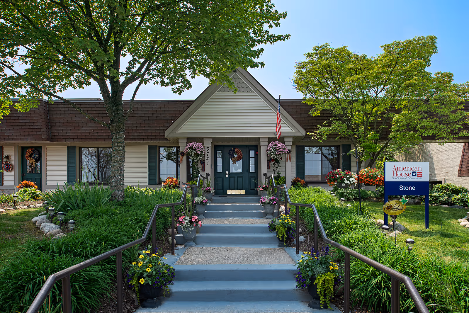 Front entrance of the American House Stone senior living building with steps, handrails, potted flowers, landscaping, and a sign.