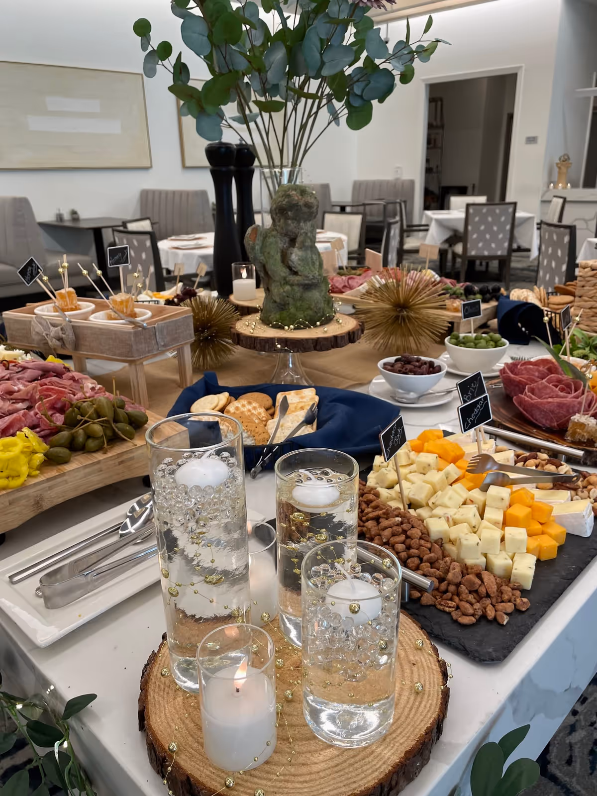 A buffet table set with a variety of cheeses, nuts, olives, crackers, and sliced meats. The table is decorated with a centerpiece featuring a small statue and greenery in a vase, along with floating candles in tall glass holders placed on wooden slices. In the background, there are dining tables and chairs in a well-lit room.