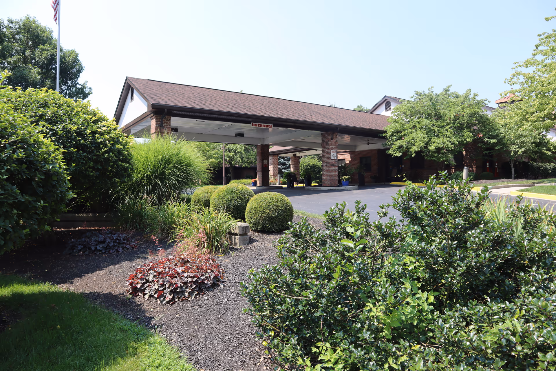 Entrance area of a senior living facility with a covered drop-off zone supported by brick pillars, surrounded by well-maintained landscaping including bushes, shrubs, and trees under a clear sky.