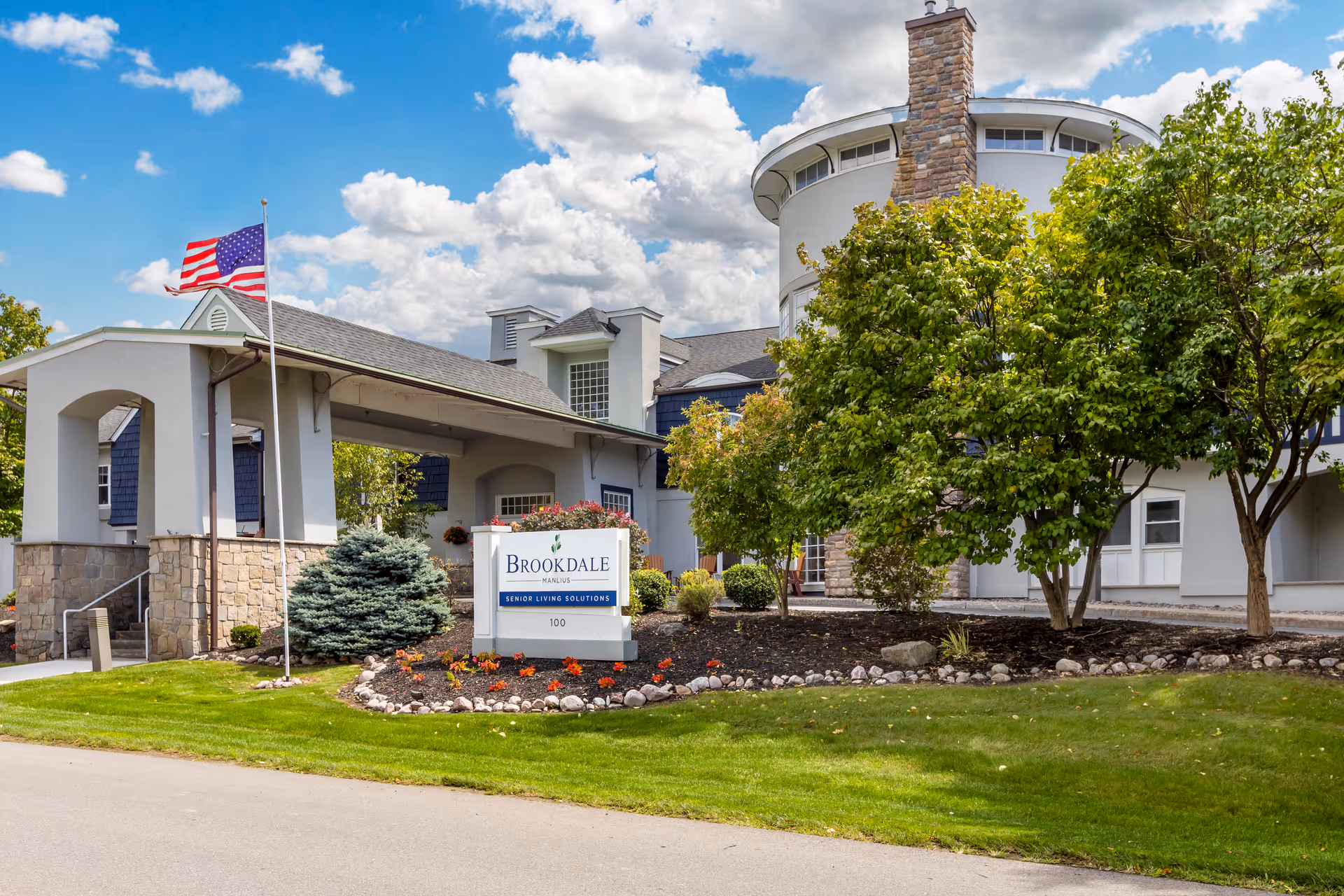 Exterior view of Brookdale Manlius senior living facility showing the entrance with a covered drop-off area, an American flag on a flagpole, landscaped greenery including trees and shrubs, and a sign reading 'Brookdale Manlius Senior Living Solutions 100' under a partly cloudy blue sky.