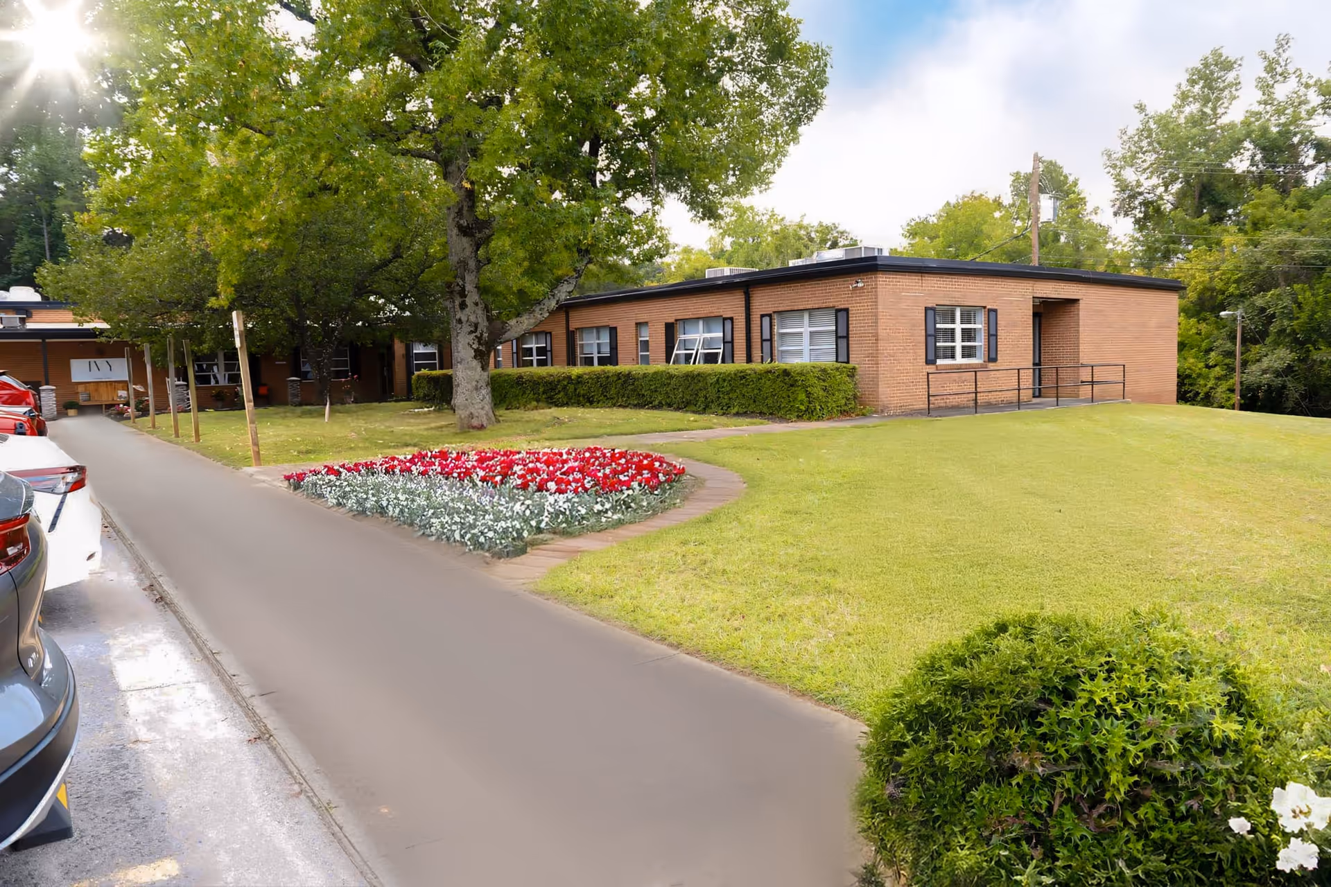 Low one-story brick building with a manicured lawn, flower bed, trees and a driveway with parked cars.