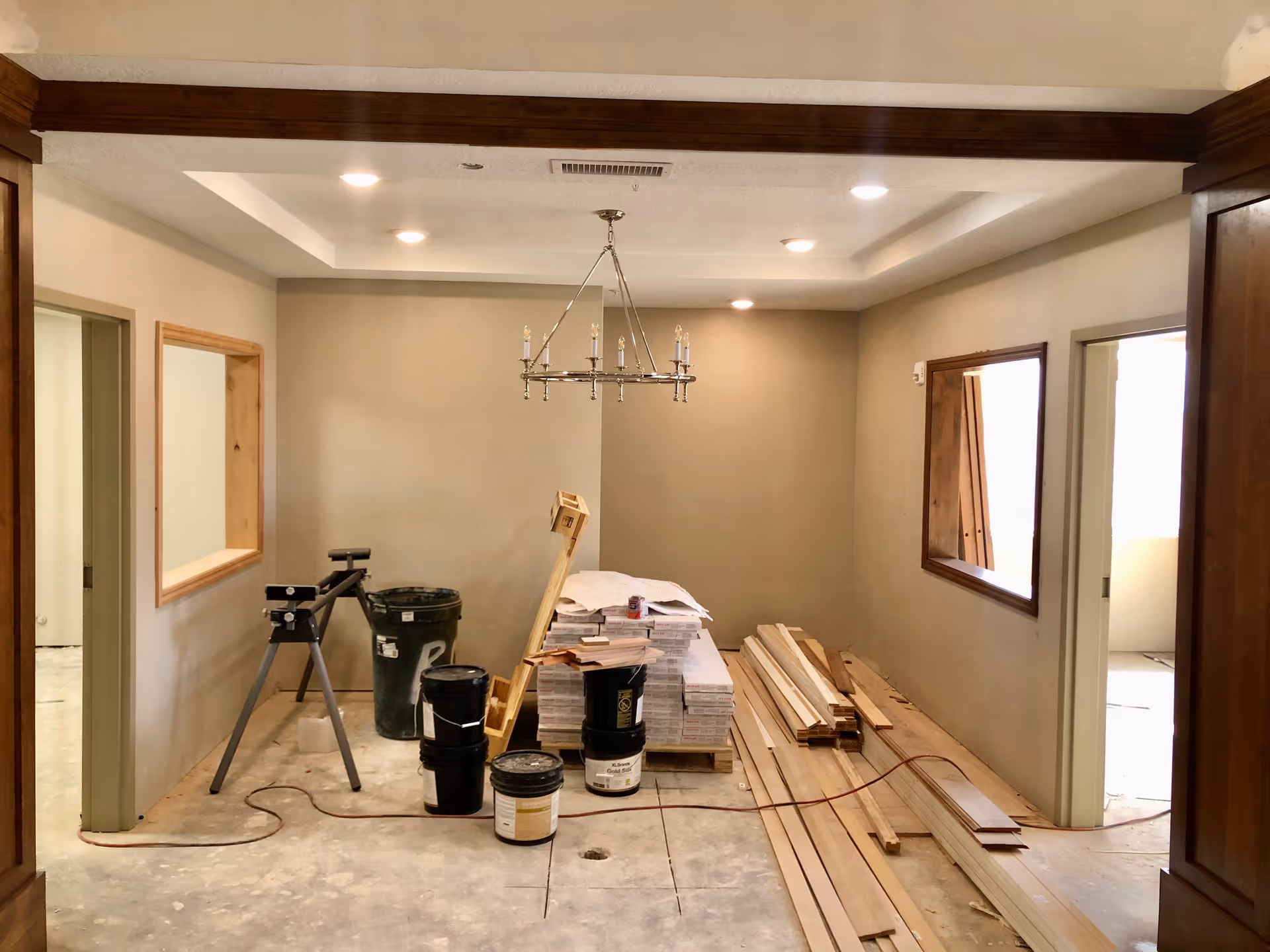 An interior room under renovation with construction materials including wooden planks, buckets, and a trash bin. The room has beige walls, recessed ceiling lights, a chandelier, and two doorways on either side. The floor is unfinished concrete.