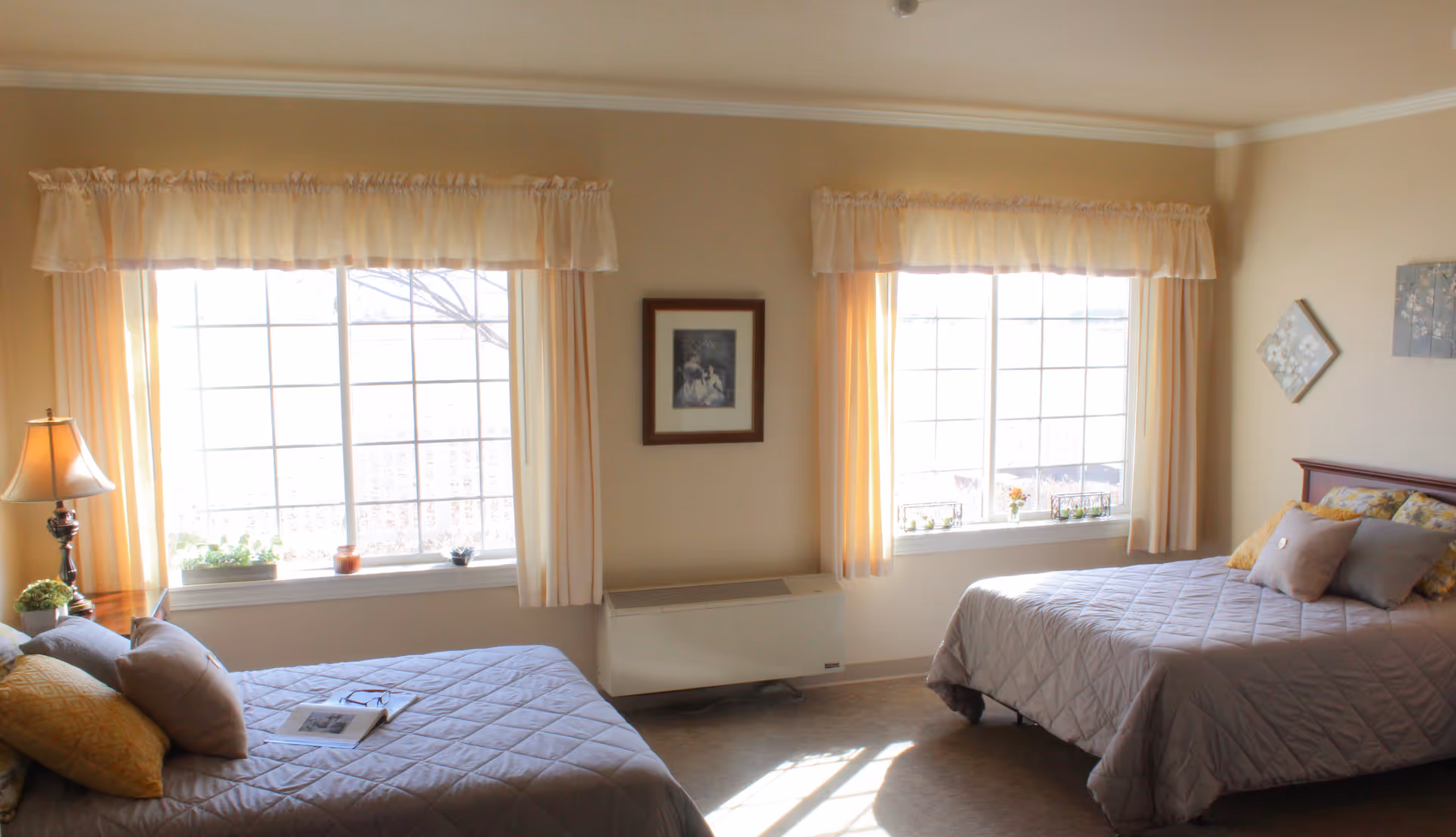 A bright bedroom with two beds covered in gray quilts and multiple pillows. There are two large windows with cream-colored curtains letting in natural light. A lamp and some small plants are on a nightstand next to one of the beds. The walls are beige with framed artwork hanging between and beside the windows.