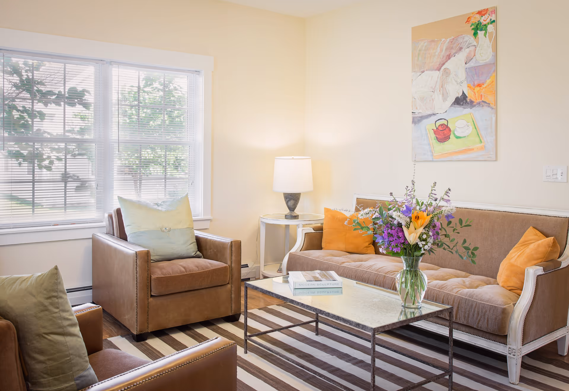 A cozy living room with a large window letting in natural light. The room features a brown sofa with orange pillows, two brown armchairs with green pillows, a glass-top coffee table with a vase of colorful flowers and books, a side table with a lamp, and a painting on the wall.