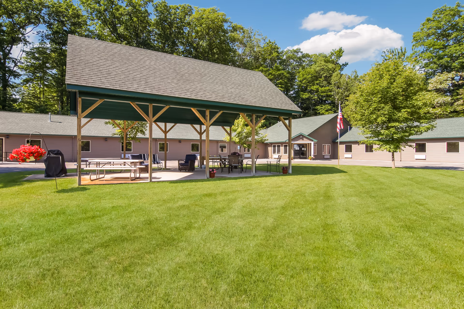 Outdoor area of South Torch Assisted Living featuring a large green lawn, a covered pavilion with tables and chairs, a barbecue grill, and a building in the background with an American flag on a flagpole. Trees surround the area under a blue sky with some clouds.
