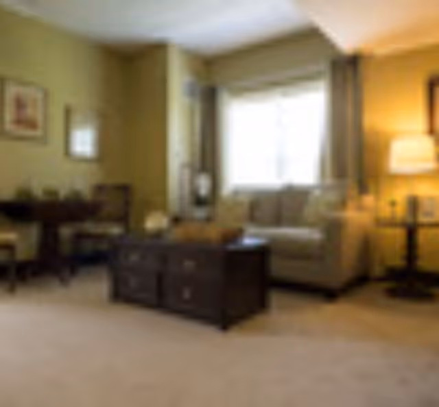 A cozy living room with a beige sofa, a dark wooden coffee table, a side table with a lamp, and a small dining area with a table and chairs against a greenish wall.
