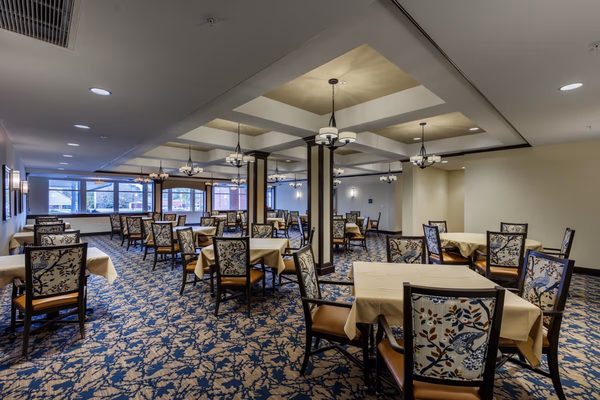 A spacious dining room with multiple tables covered in beige tablecloths and surrounded by chairs with floral patterned upholstery. The room features a blue and beige patterned carpet, large windows letting in natural light, and ceiling lights with chandeliers hanging from a coffered ceiling.