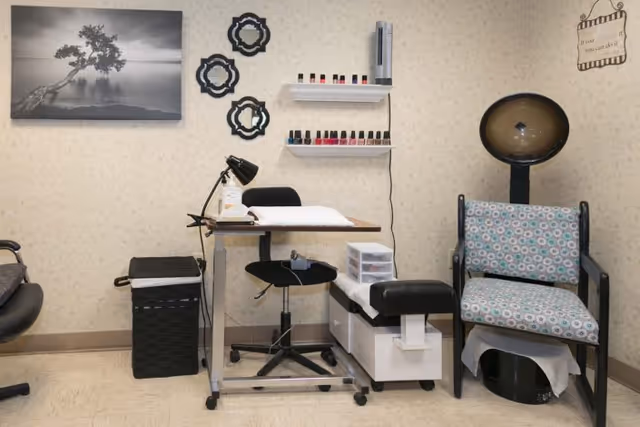 Small salon area with a manicure table, nail polish shelves, a client chair under a hooded dryer, and wall decorations.