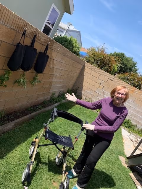 An elderly woman with glasses and a purple long-sleeve shirt stands on a grassy backyard next to a walker. She is smiling and gesturing with one hand towards the walker. Behind her is a brick wall with three black hanging planters and some plants growing in the garden bed. The sky is clear and blue.