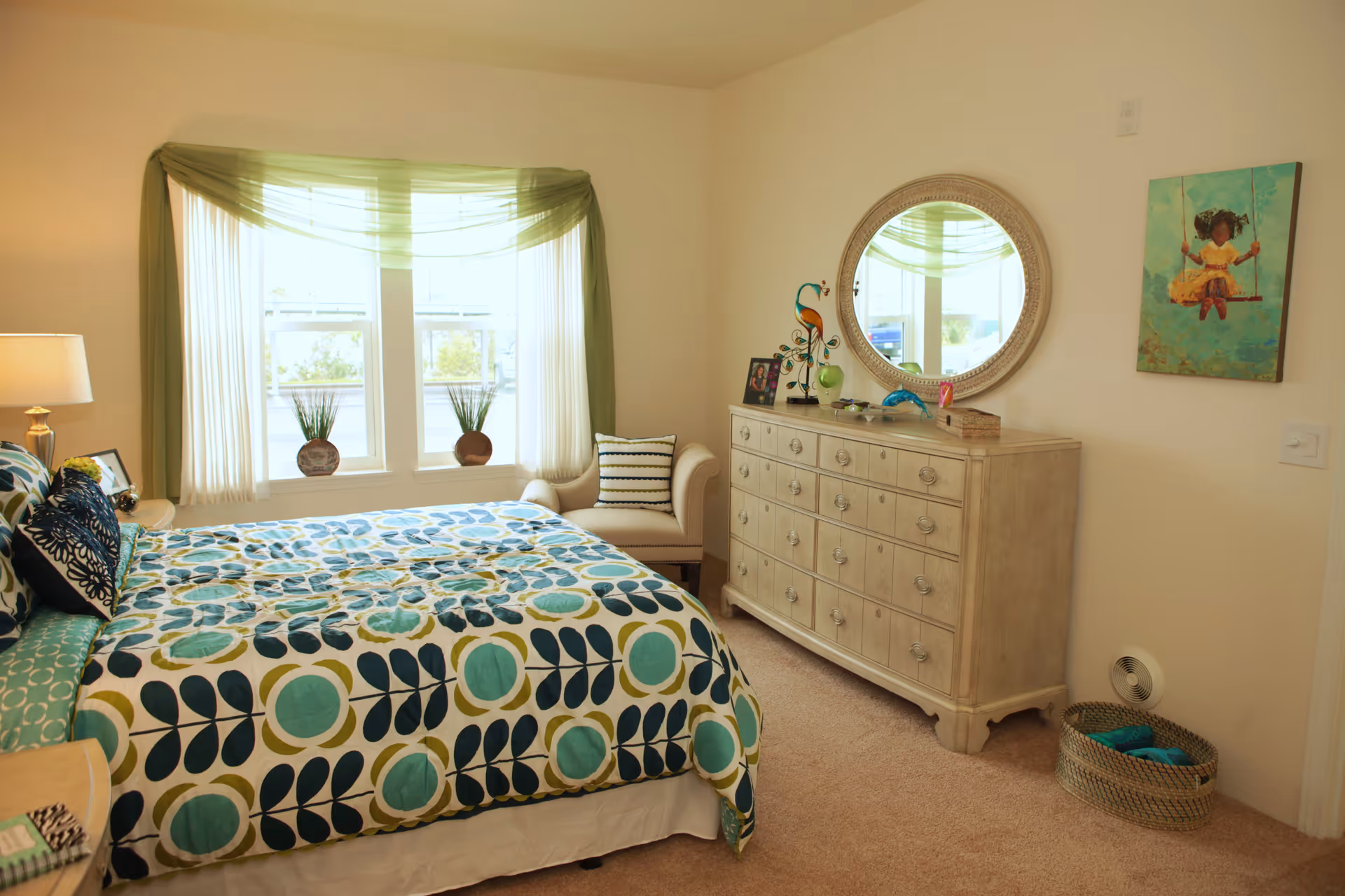 A bright bedroom with a large bed covered in a colorful patterned bedspread in shades of blue, green, and white. There is a window with sheer white curtains and a green valance, two potted plants on the windowsill, a beige armchair with a striped pillow, a light-colored wooden dresser with multiple drawers, a round mirror above the dresser, and a painting of a child on a swing on the wall. A woven basket with blue towels is on the floor near the dresser.