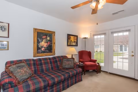 A cozy living room with a plaid sofa adorned with two decorative pillows, a red upholstered armchair, a floor lamp, and framed artwork on the walls. French doors with glass panes open to a grassy outdoor area with buildings visible in the background.