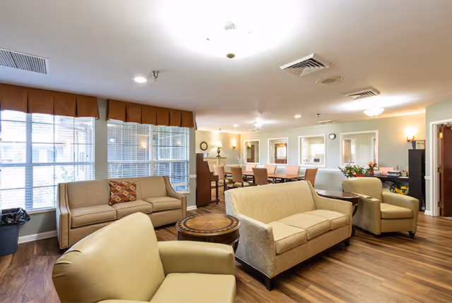 A bright and spacious senior living facility common area with multiple seating options including beige sofas and armchairs arranged around a round wooden coffee table. Large windows with brown valances allow natural light to fill the room. In the background, there is a dining area with a long table and chairs, and soft wall lighting creates a warm atmosphere.