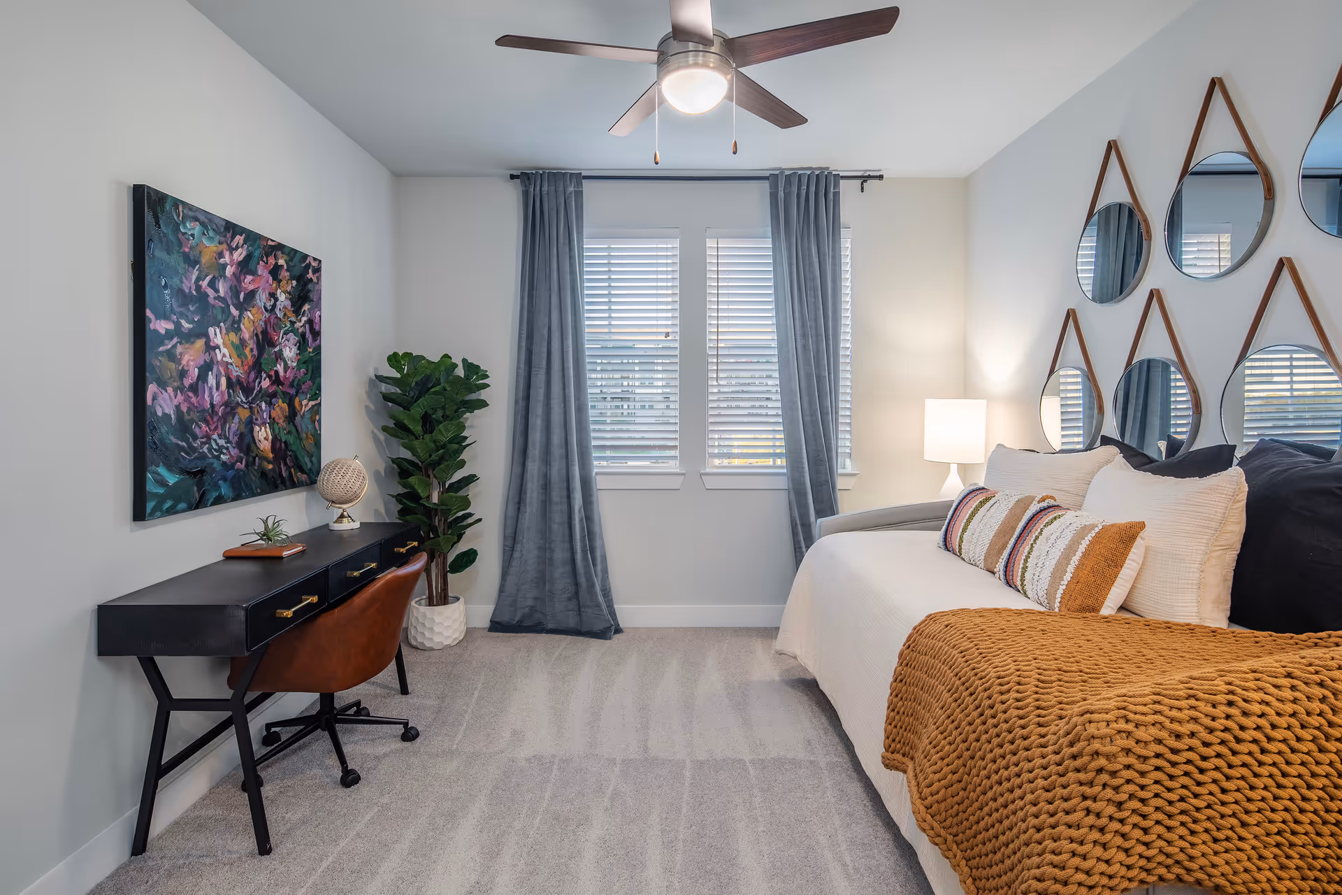 A cozy bedroom with a daybed covered in white bedding and a mustard-colored knitted throw blanket. The bed has several decorative pillows. Above the bed, there are six round mirrors with leather straps arranged in two rows on the wall. To the left, there is a black desk with three drawers, a brown leather swivel chair, a small globe, and a plant on the desk. A large colorful abstract painting hangs on the wall above the desk. Two windows with white blinds and gray curtains are centered on the back wall. A ceiling fan with a light is mounted on the ceiling, and a white table lamp is on a small side table next to the bed. The floor is carpeted in a light gray color.