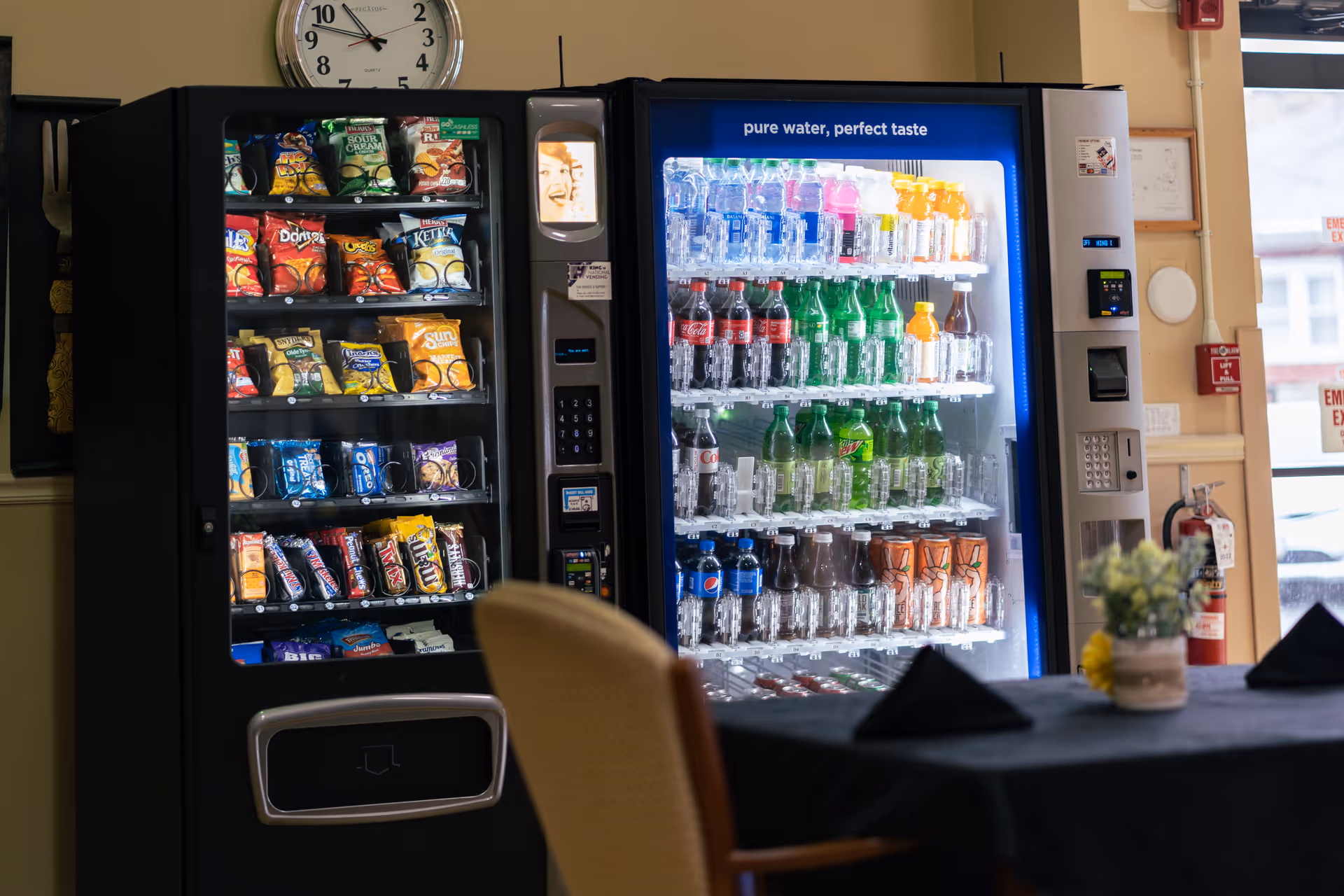 Two vending machines side by side indoors, one stocked with various snacks like chips and candy bars, the other stocked with bottled drinks including water, soda, and juice. A clock is mounted on the wall above the vending machines. In the foreground, there is a table with a black tablecloth, a small flower arrangement, and folded black napkins. A window and emergency exit signs are visible in the background.