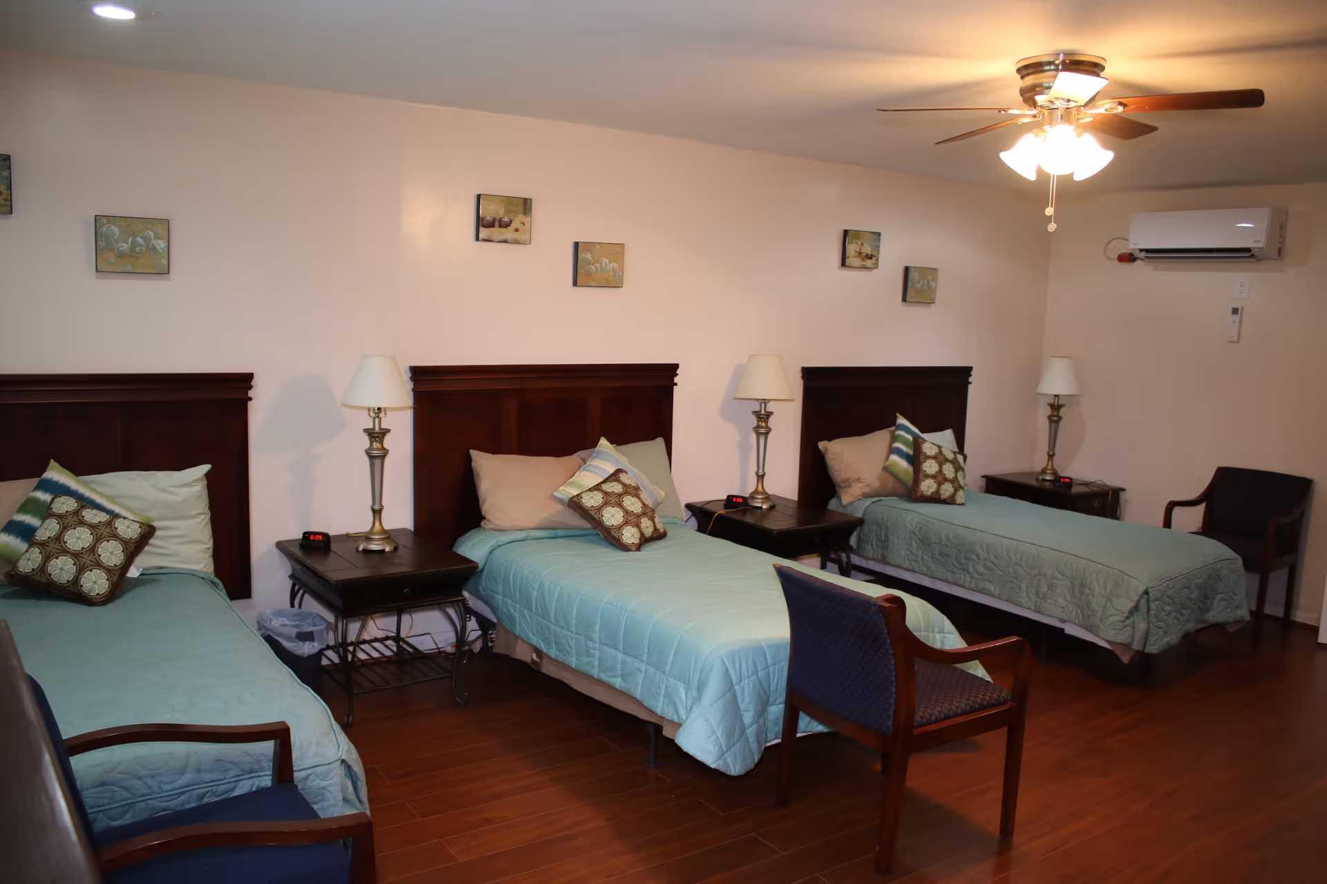 A bedroom with three single beds arranged side by side, each with a wooden headboard, light blue bedspreads, and decorative pillows. Between the beds are two nightstands with lamps and alarm clocks. The room has wooden flooring, a ceiling fan with lights, and a wall-mounted air conditioning unit. There are small framed pictures on the wall above the beds and two chairs placed near the beds.