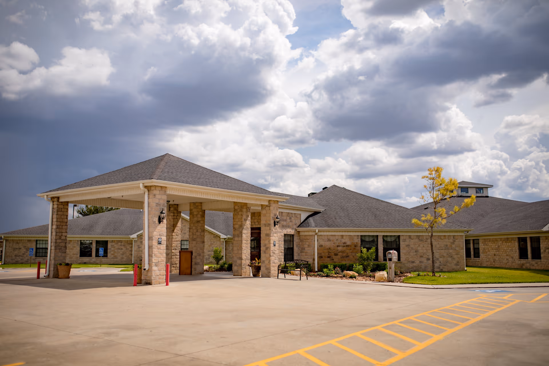 Front exterior of a low-rise brick nursing facility with a covered porte-cochere entrance under a cloudy sky.