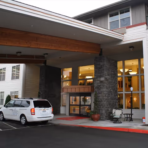 Entrance of Bonaventure of East Wenatchee senior living facility showing a covered drop-off area with stone pillars, large windows, a white van parked nearby, and outdoor seating with a table and chairs.